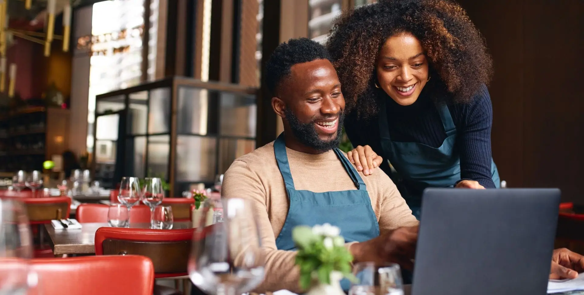 Two restaurant owners smiling as they review results on a laptop in their dining room